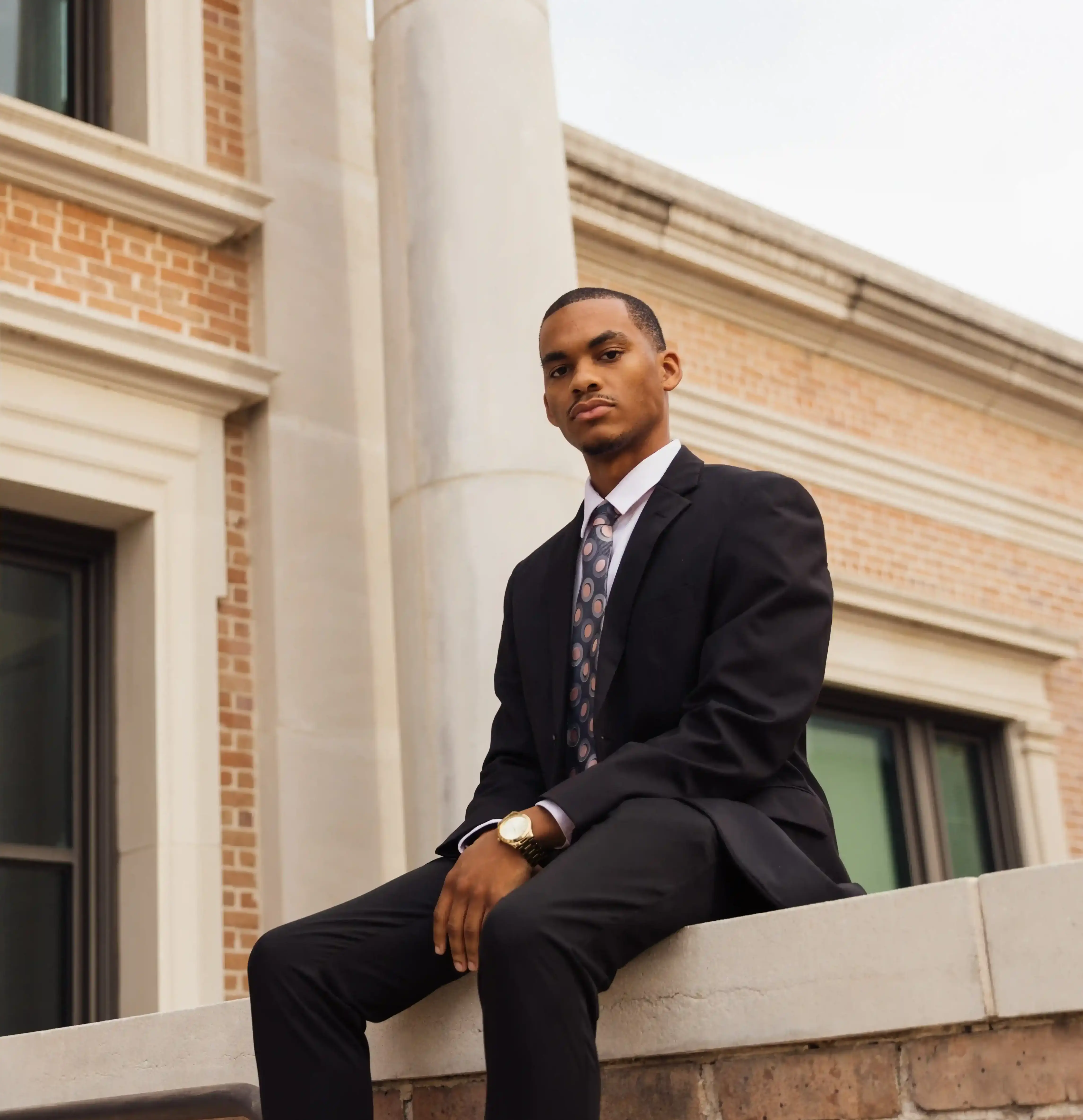 a young, handsome, black man in a black suit sitting atop a ledge in front of a building