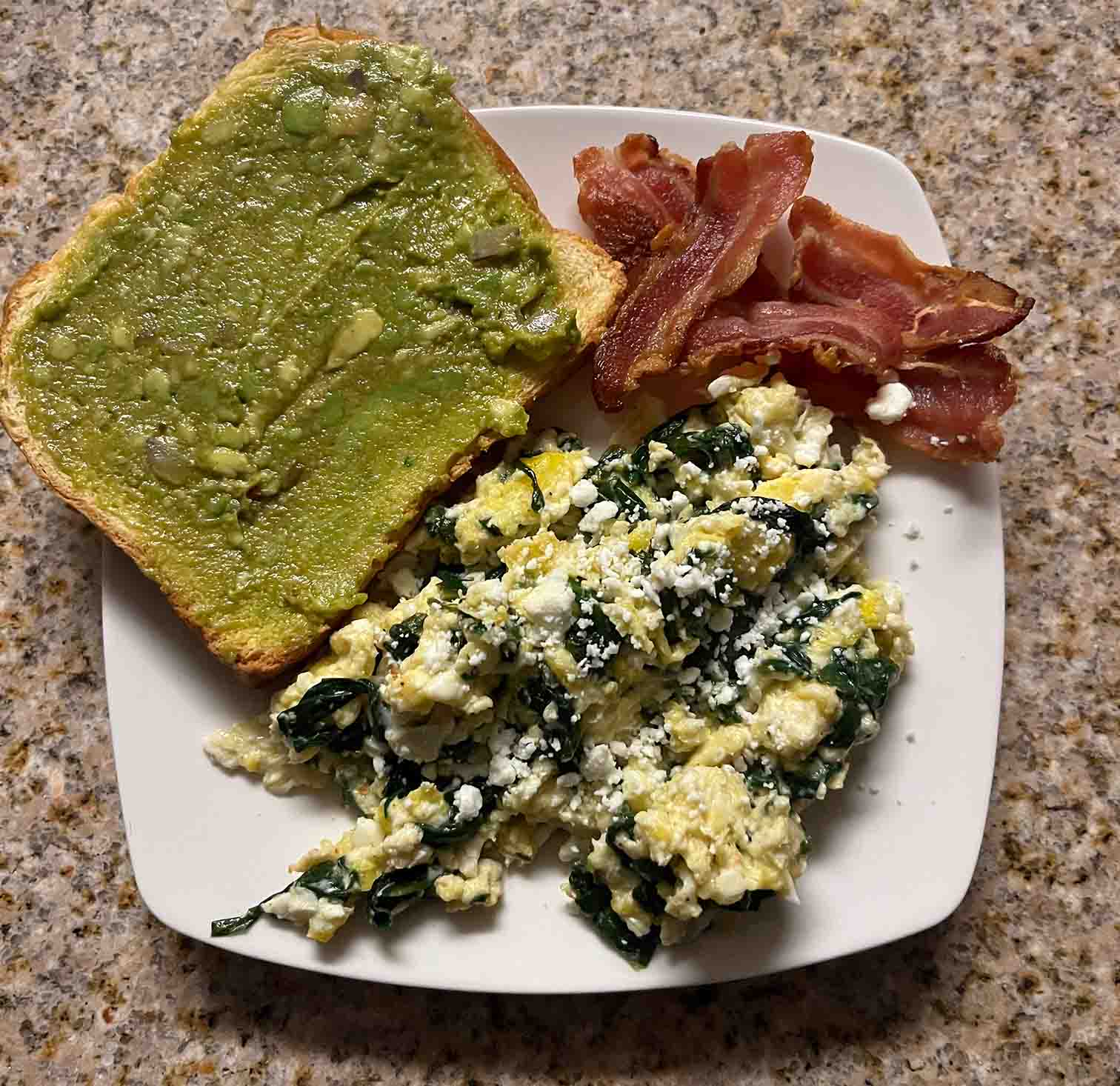 a white plate containing scrambled eggs with spinach, slices of bacon, and a piece of toast with a guacamole spread