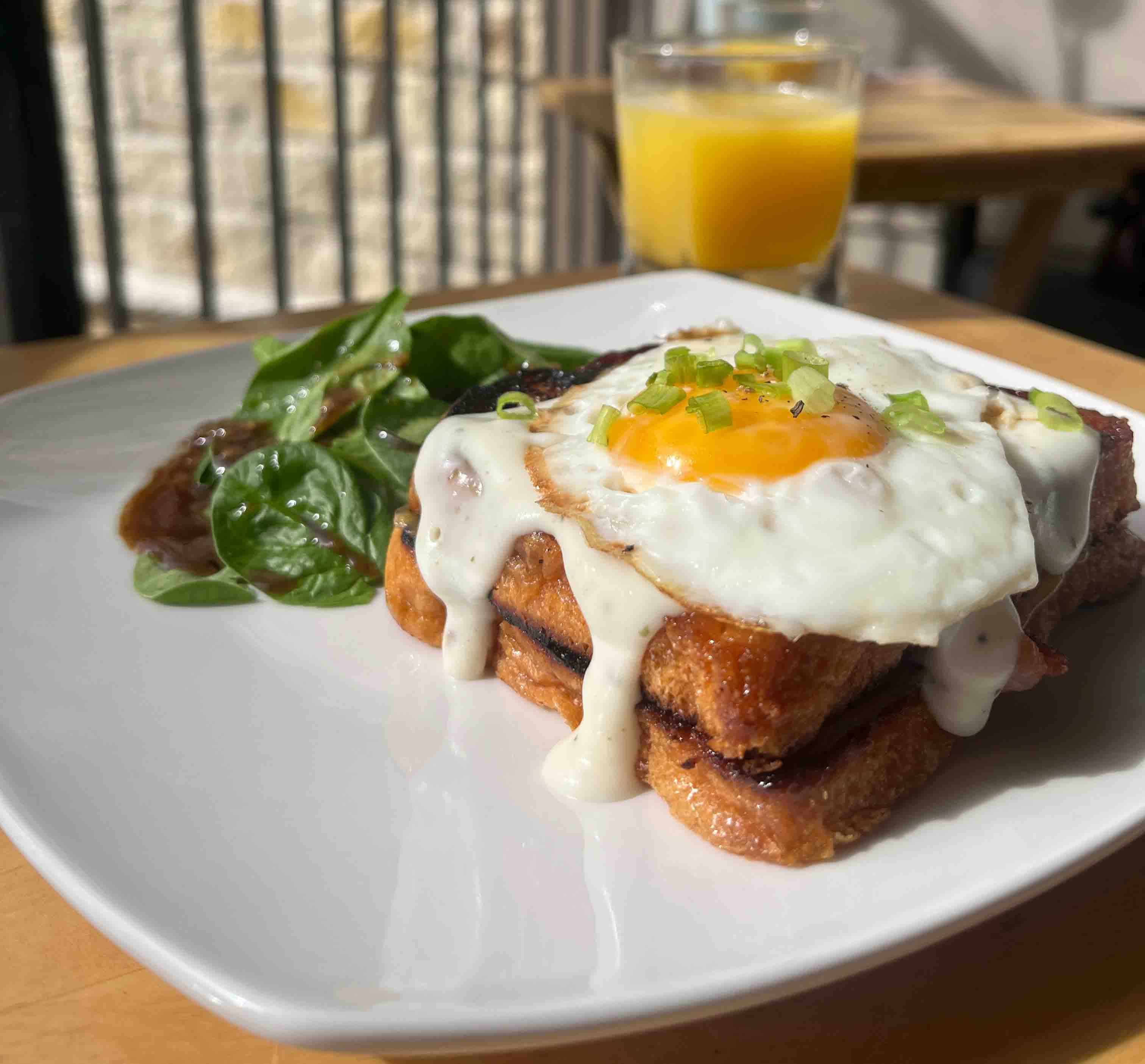 a croque madame sandwich, topped with an over-easy egg, served with leaves of spinach and a balsamic vinaigrette dressing, with a glass of orange juice