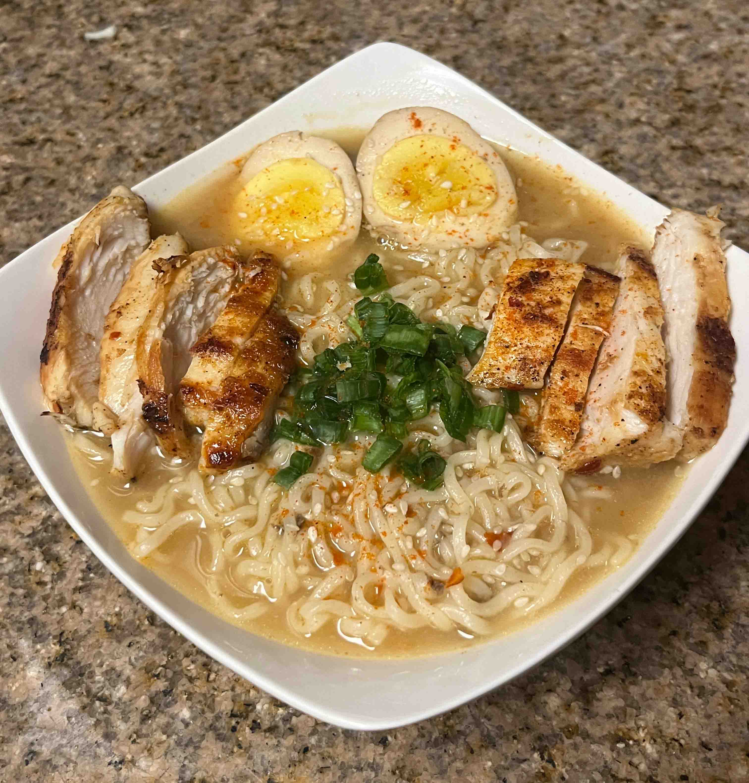 ramen noodles in a white bowl, with sliced chicken, green onions, and a boiled egg sliced in half