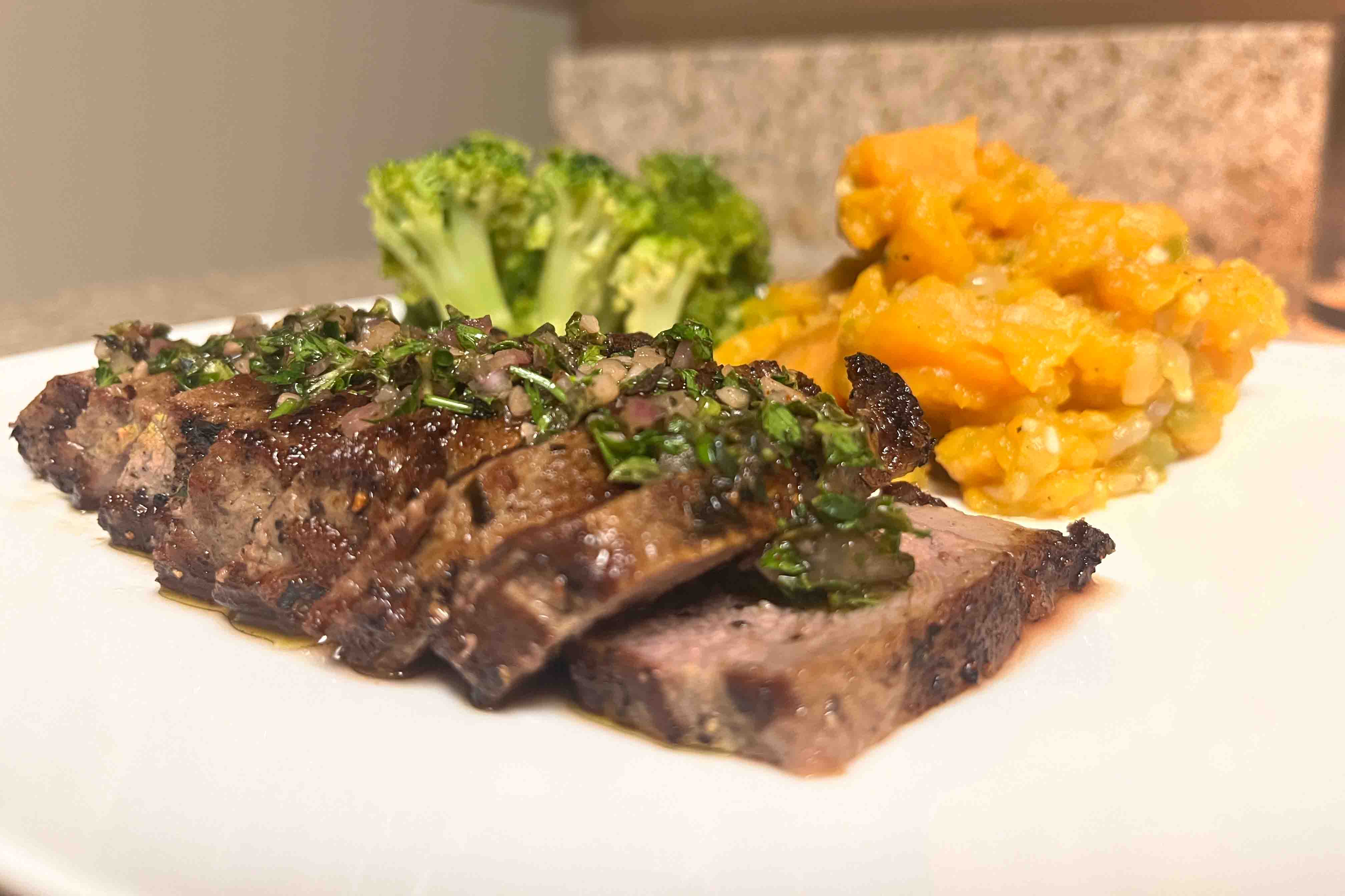 sliced steak topped with chimichurri, with broccoli and sweet potatoes behind it